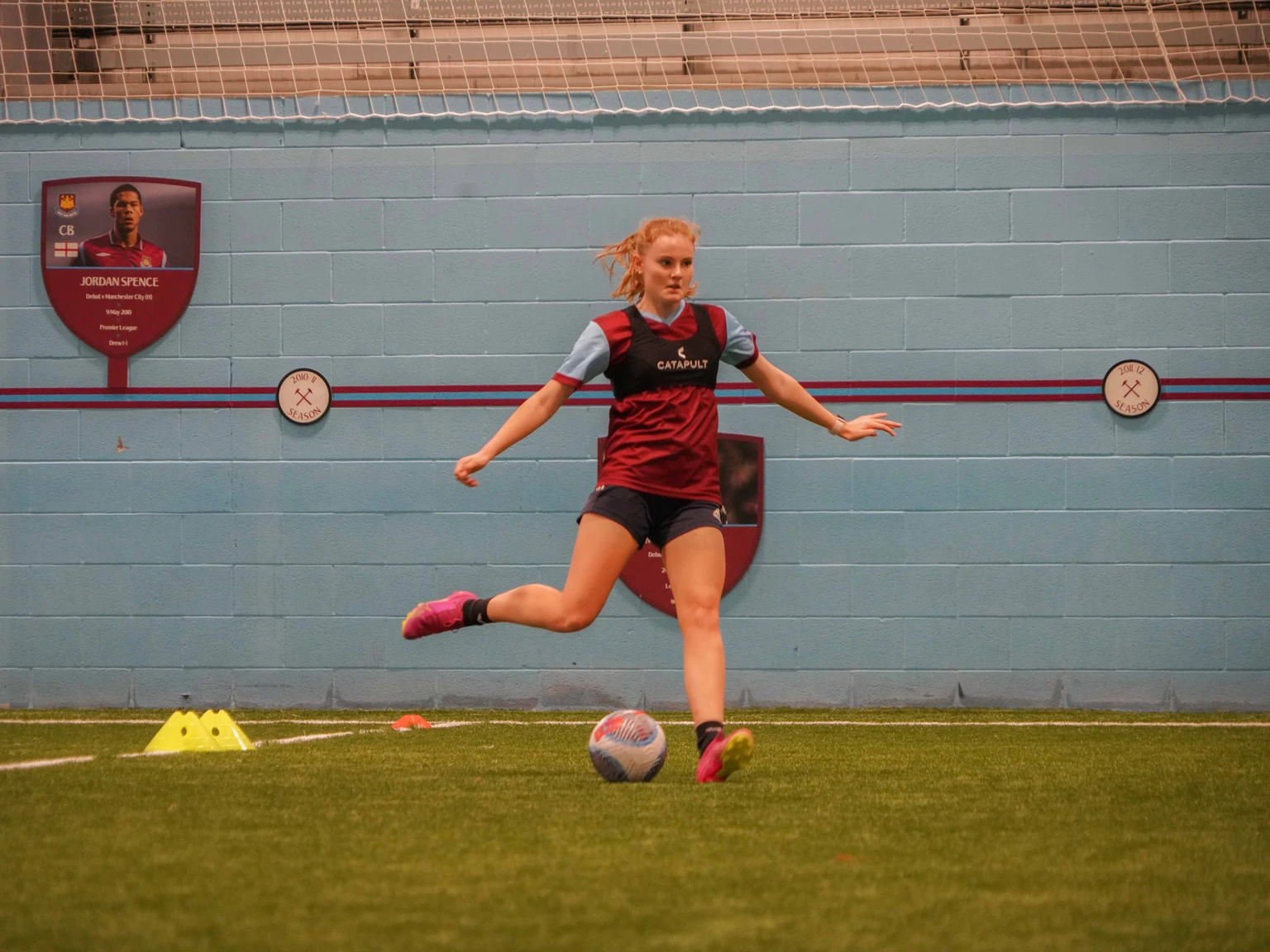 Victoria striking a football during training on an indoor pitch.